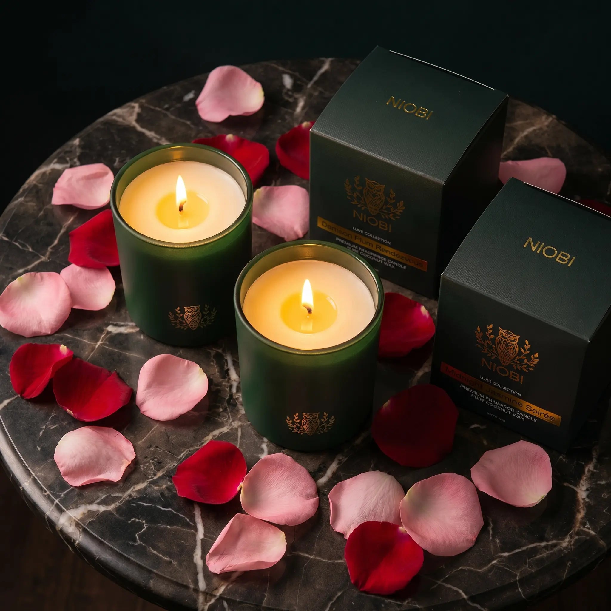 NIOBI Ритуал за двама две запалени ароматни свещи с кутии Damson Plum Rendezvous и Midnight Jasmine Soirée върху тъмен мрамор с розови листенца
