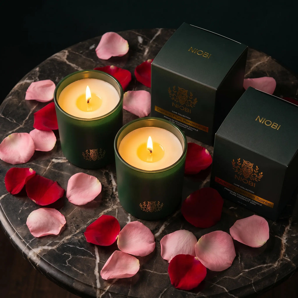NIOBI Ритуал за двама две запалени ароматни свещи с кутии Damson Plum Rendezvous и Midnight Jasmine Soirée върху тъмен мрамор с розови листенца
